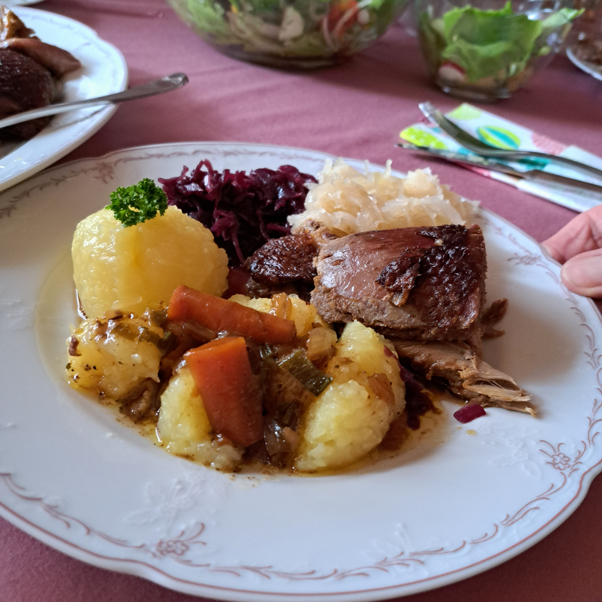 A plate featuring a succulent piece of roasted duck, a sliced potato dumpling bathed in rich gravy, and a served with blaukraut and sauerkraut.