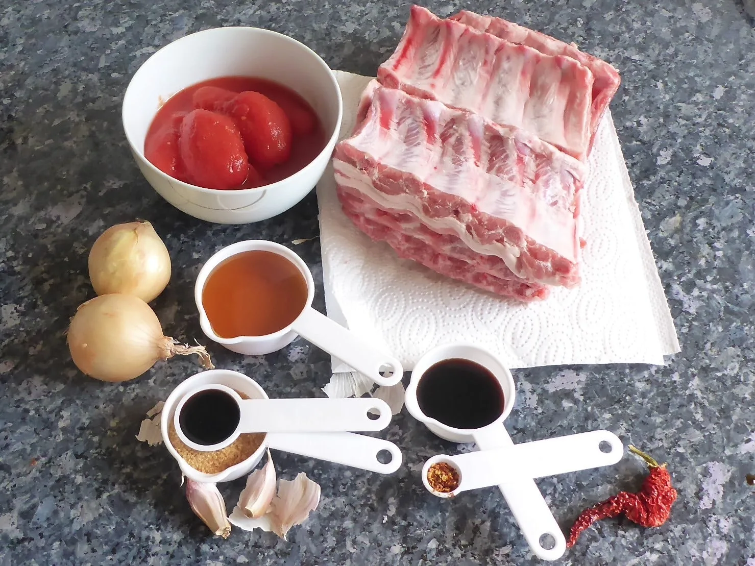 A neatly arranged assortment of ingredients for homemade BBQ pork spare ribs, including raw pork spare ribs, canned tomatoes, soy sauce, vinegar, sugar, garlic, and various spices on a kitchen countertop.