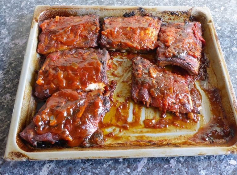 Freshly baked pork spare ribs on a baking sheet, glistening with a rich homemade BBQ sauce, with a caramelized crust and tender, juicy texture.