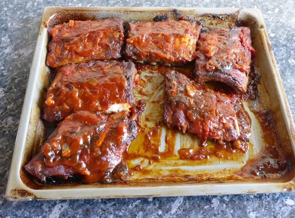 Freshly baked pork spare ribs on a baking sheet, glistening with a rich homemade BBQ sauce, with a caramelized crust and tender, juicy texture.