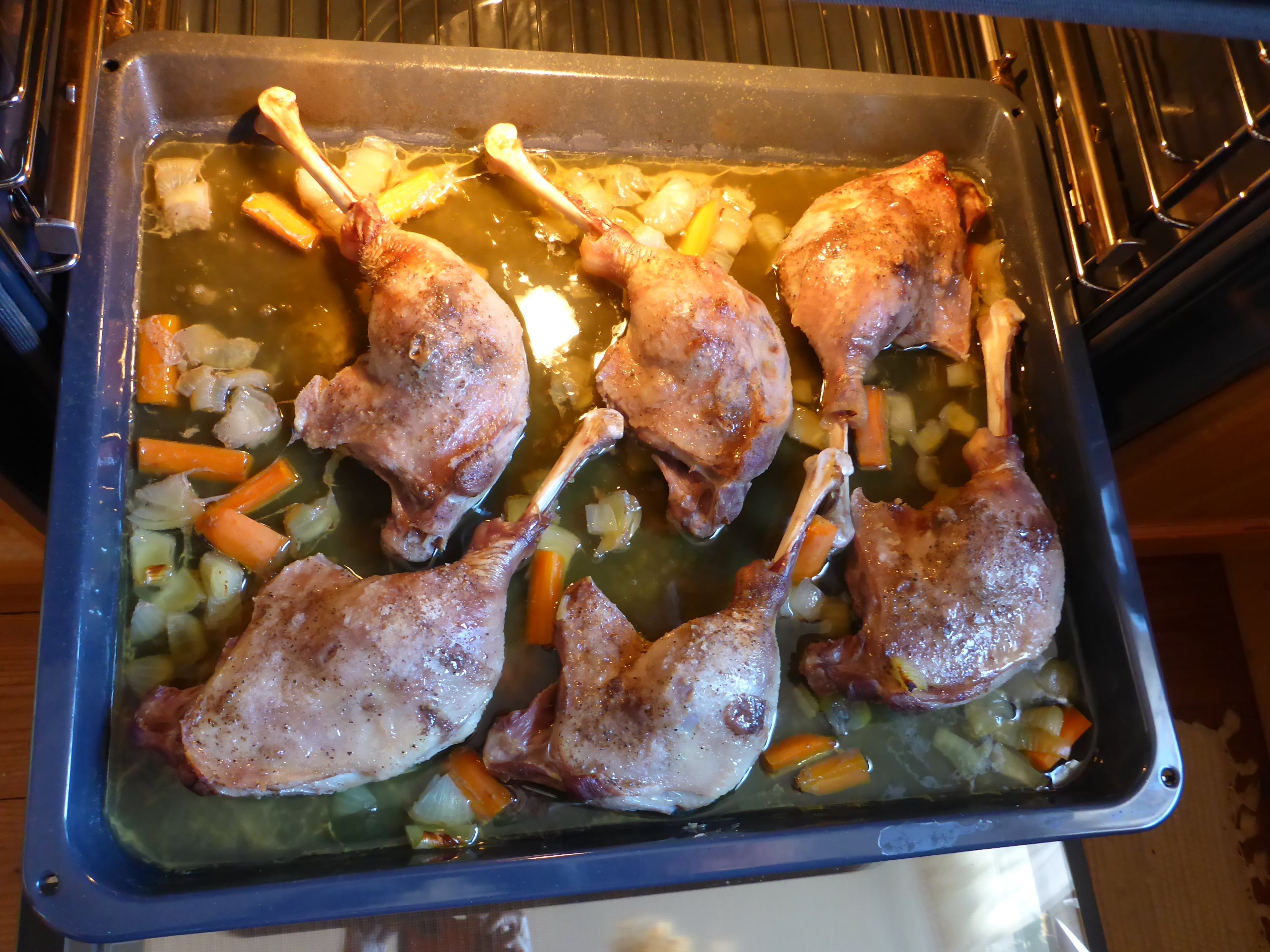 Goose legs and diced carrots and onions on a baking sheet inside the oven, midway through the roasting process.