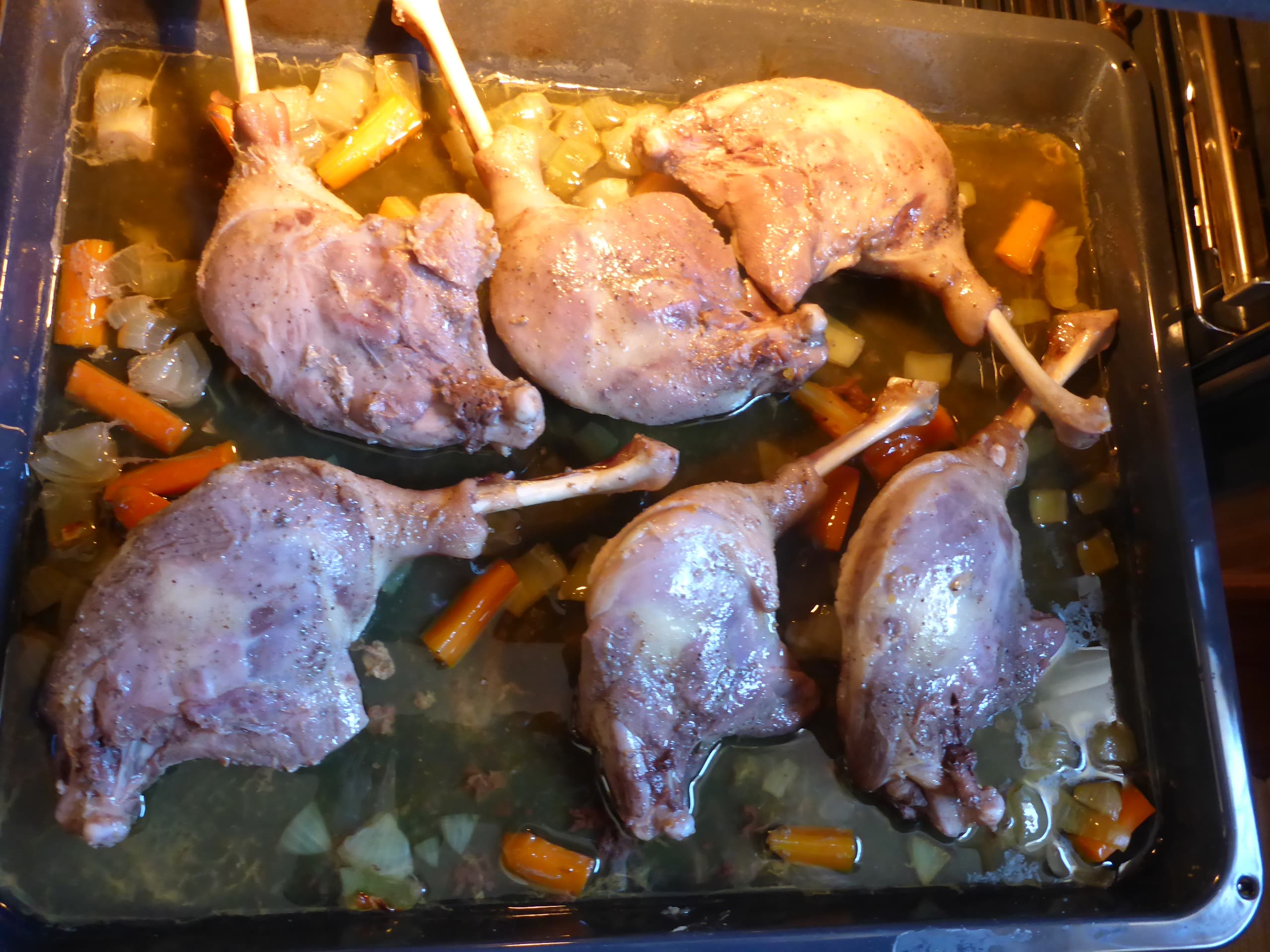 Goose legs and diced carrots and onions on a baking sheet inside the oven, midway through the roasting process.