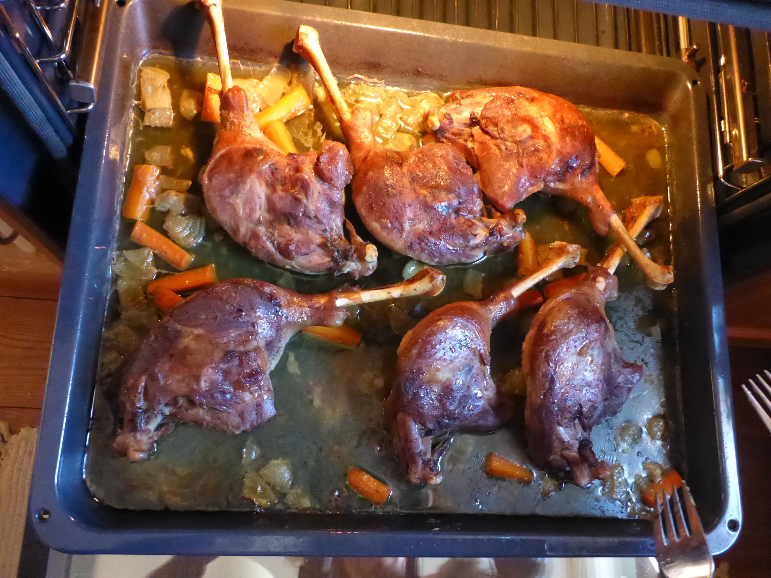 Goose legs and diced carrots and onions on a baking sheet inside the oven, almost through with the roasting process.