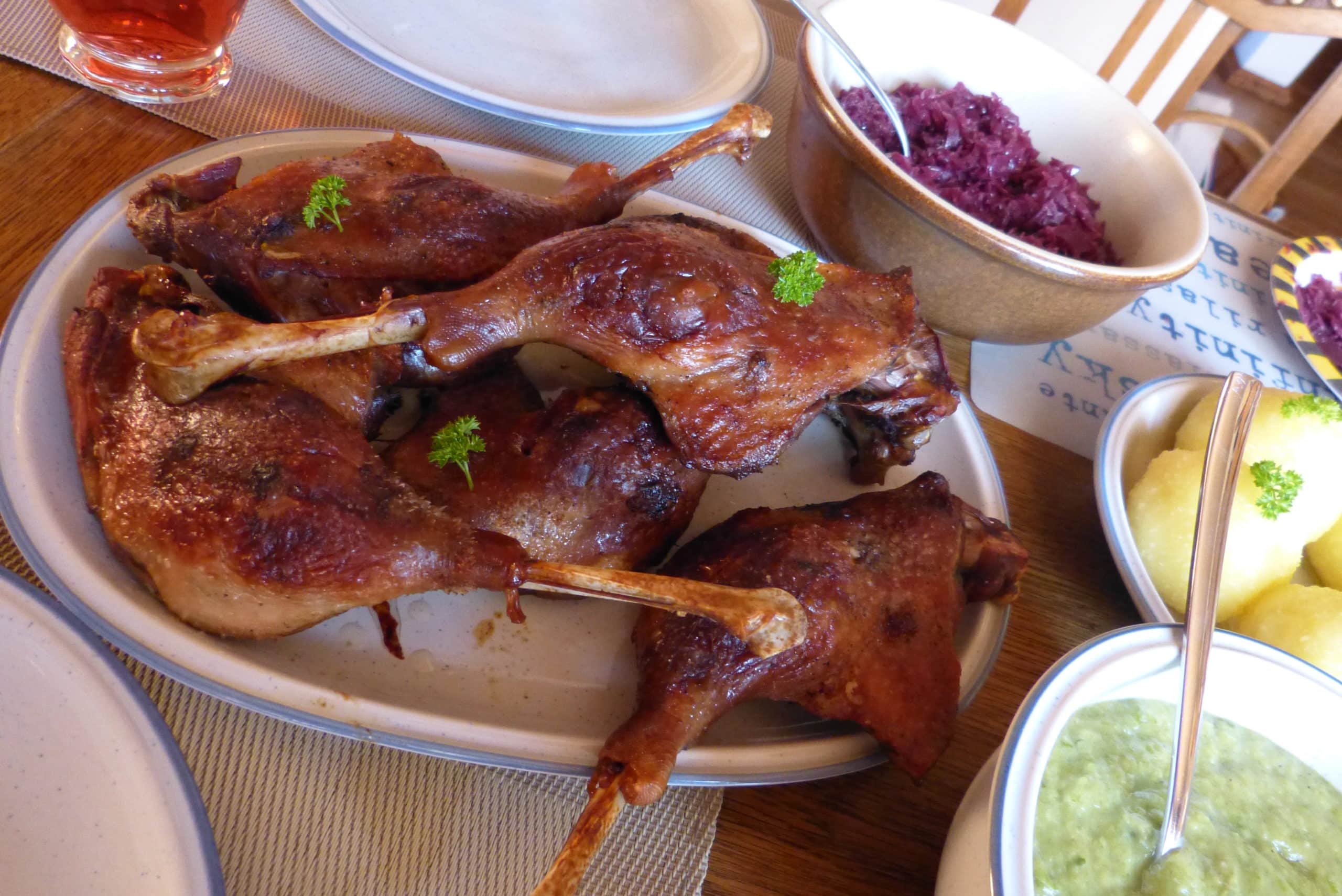Beautifully golden brown German roasted goose legs on a platter, accompanied by a plate of potato dumplings, a bowl of savoy cabbage (Wirsing), and a bowl of blaukraut.