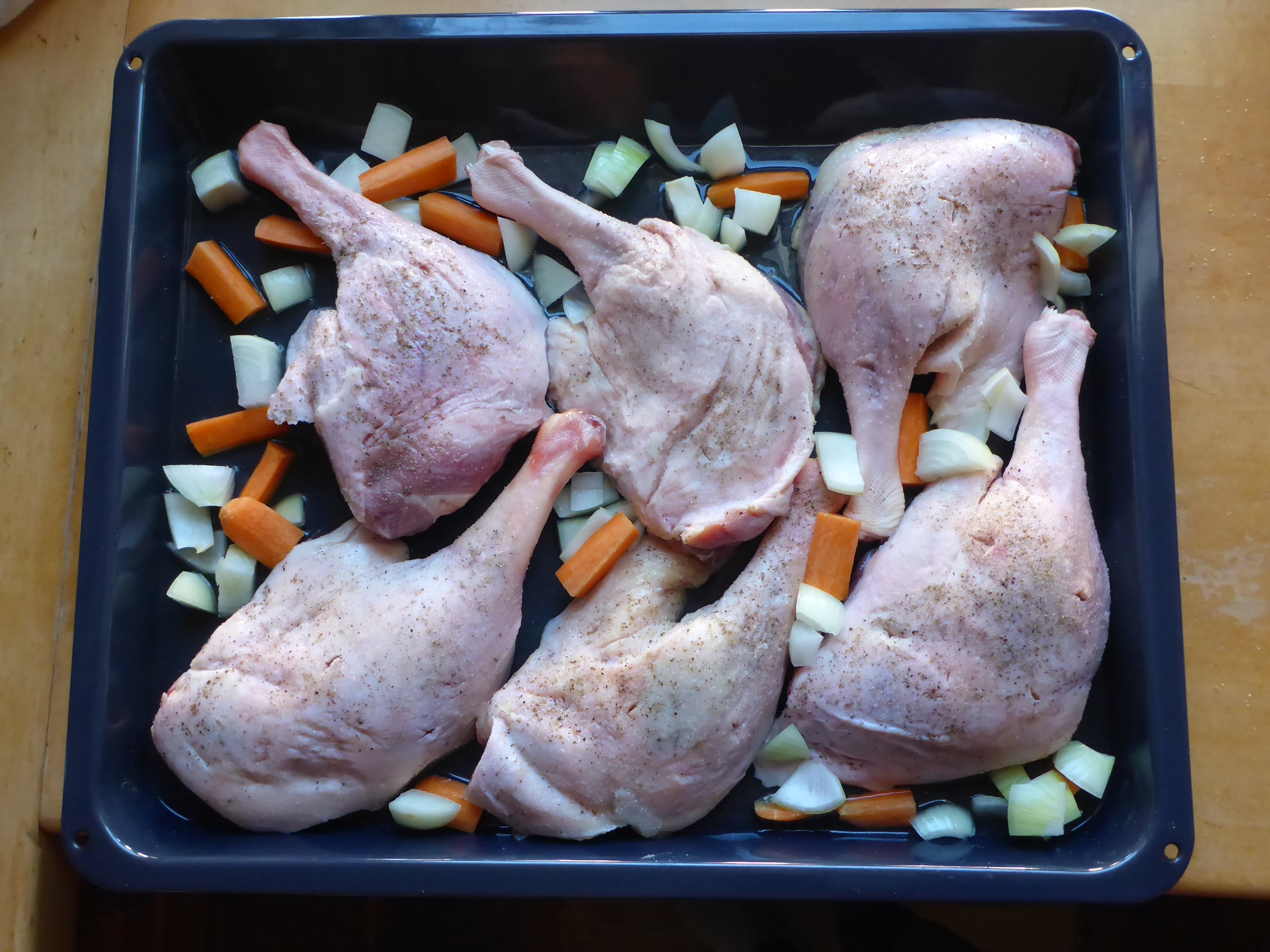 Goose legs on a baking sheet surrounded by small pieces of carrots and onions, ready to be placed in the oven for roasting.