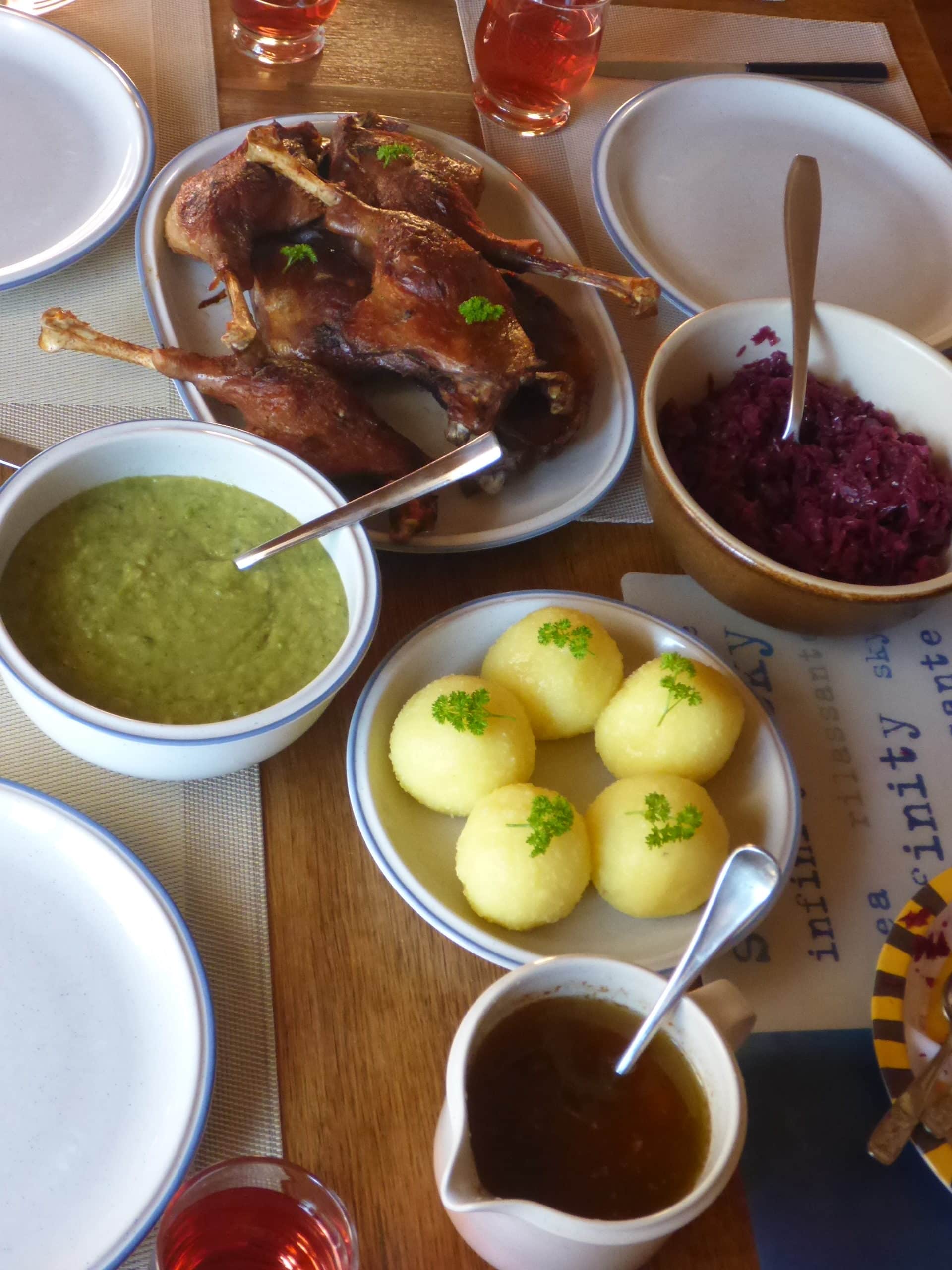 Sunday lunch in Bavaria: A photo featuring perfectly golden brown German roasted goose legs on a platter, accompanied by a plate of potato dumplings, a bowl of savoy cabbage (Wirsing), gravy,  and a bowl of blaukraut, all elegantly served on a dining table.