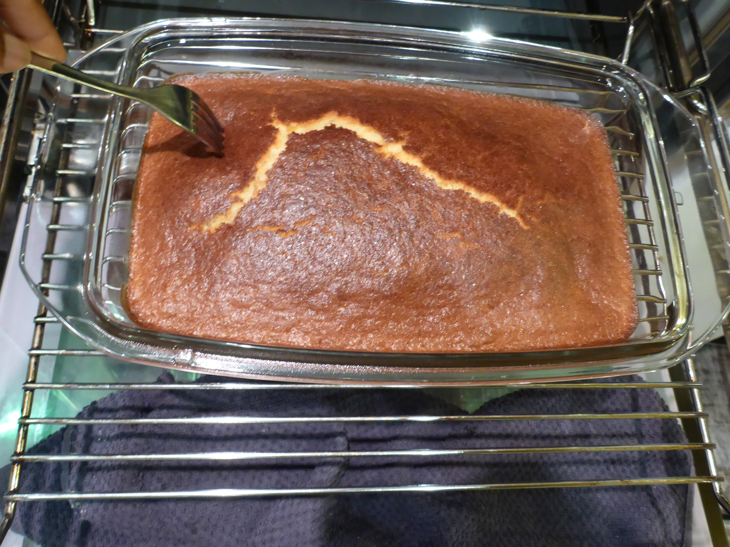 Ein Bild von einem frisch gebackenen Malva Pudding, der gerade aus dem Ofen genommen wurde und mit einer Gabel gepickst wird, um sicherzustellen, dass er den Sirup gut aufsaugt.