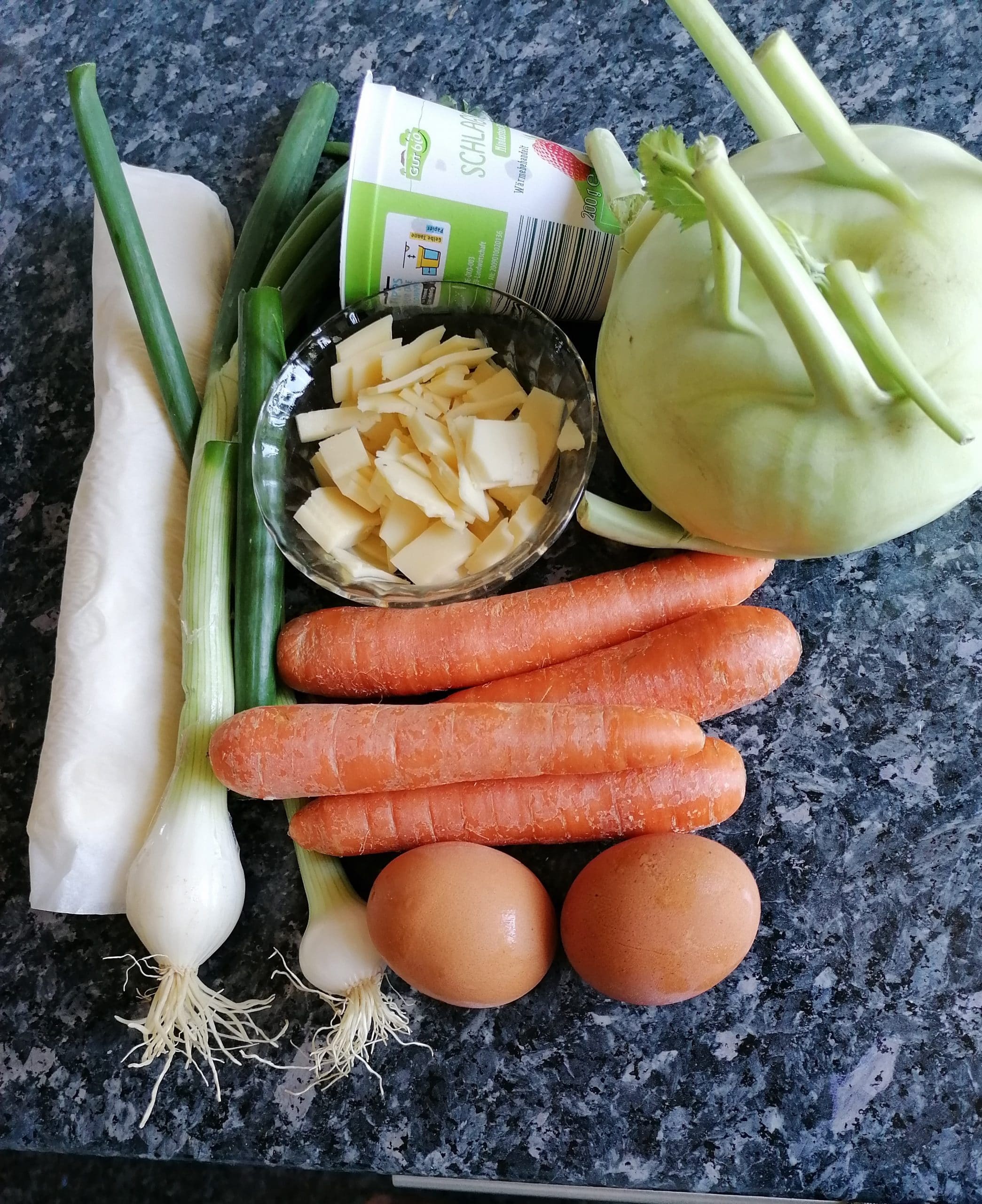 A vibrant photo showcasing the fresh ingredients for making Kohlrabi Quiche: kohlrabi bulbs, carrots, spring onions, Gouda cheese, eggs, heavy cream, and store-bought puff pastry arranged on a kitchen counter. A delightful and wholesome combination for crafting a delicious quiche.