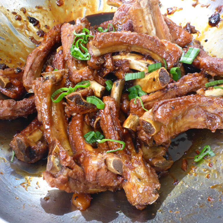 Ein Wok mit glänzend glasierten süß-sauren Rippchen, garniert mit frisch geschnittenen Frühlingszwiebeln.