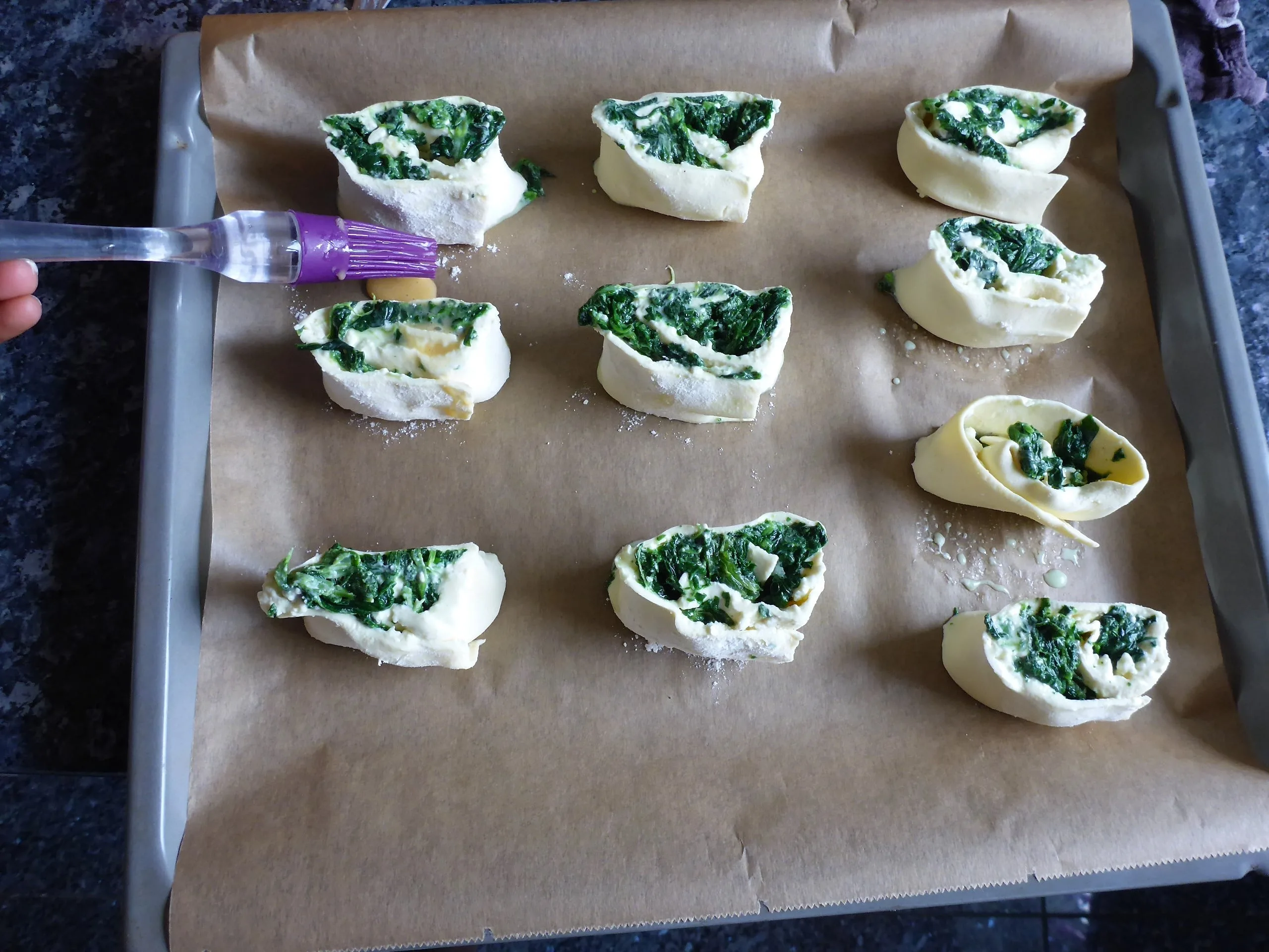 Auf dem Bild sind Spinat und Feta Blätterteigschnecken vegetarisch vor dem Backen, die mit einer Ei-Milch Mischung bestrichen werden, zu sehen.