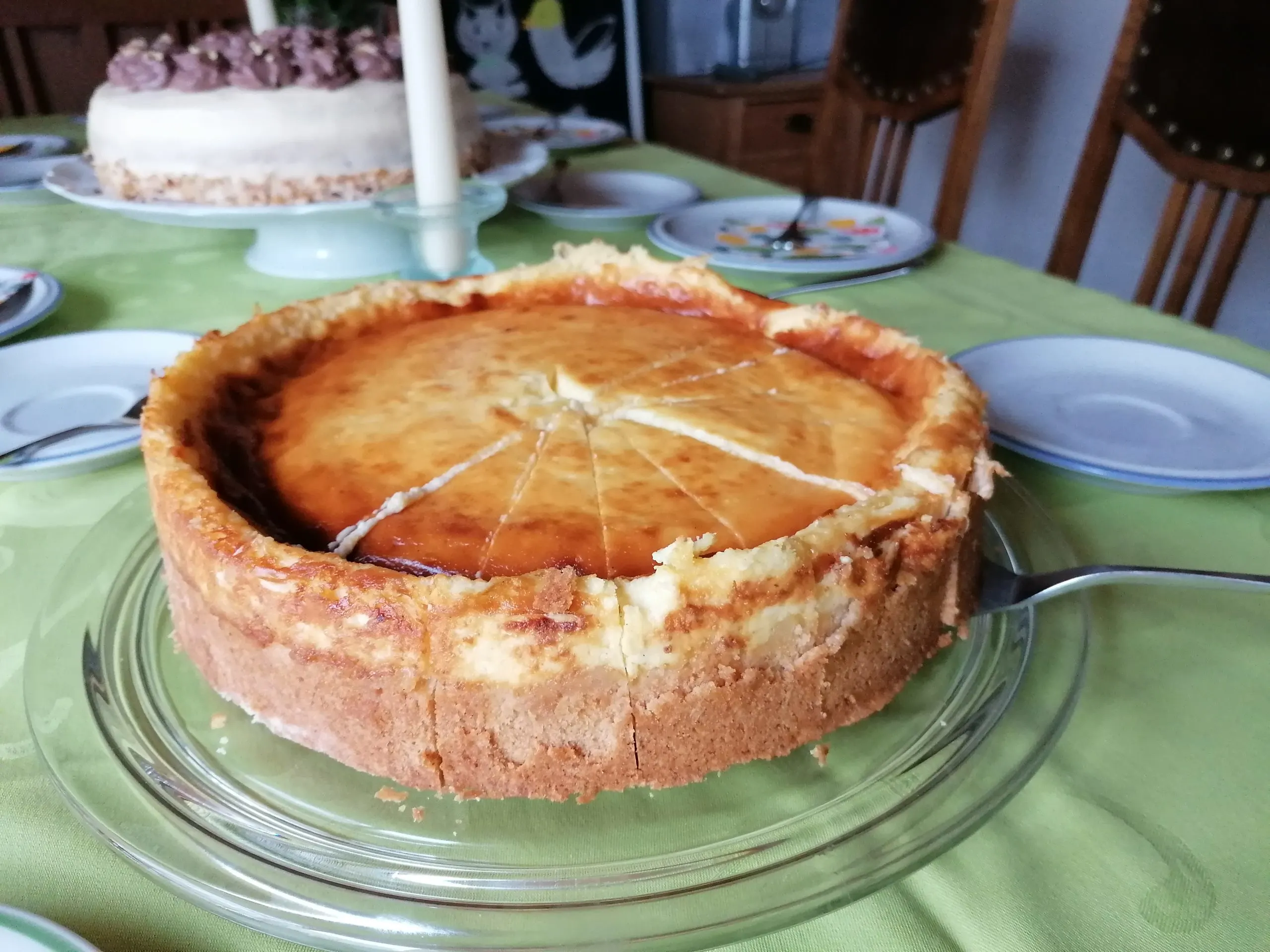 Ein festlich gedeckter Tisch für einen Geburtstagsmittagskaffee, auf dem ein köstlicher Käsekuchen ohne Quark mit einer wunderschönen dunklen Oberfläche präsentiert wird. Neben dem Käsekuchen steht verlockend eine Buttercremetorte, beide Leckereien bereit, um den besonderen Anlass zu feiern.