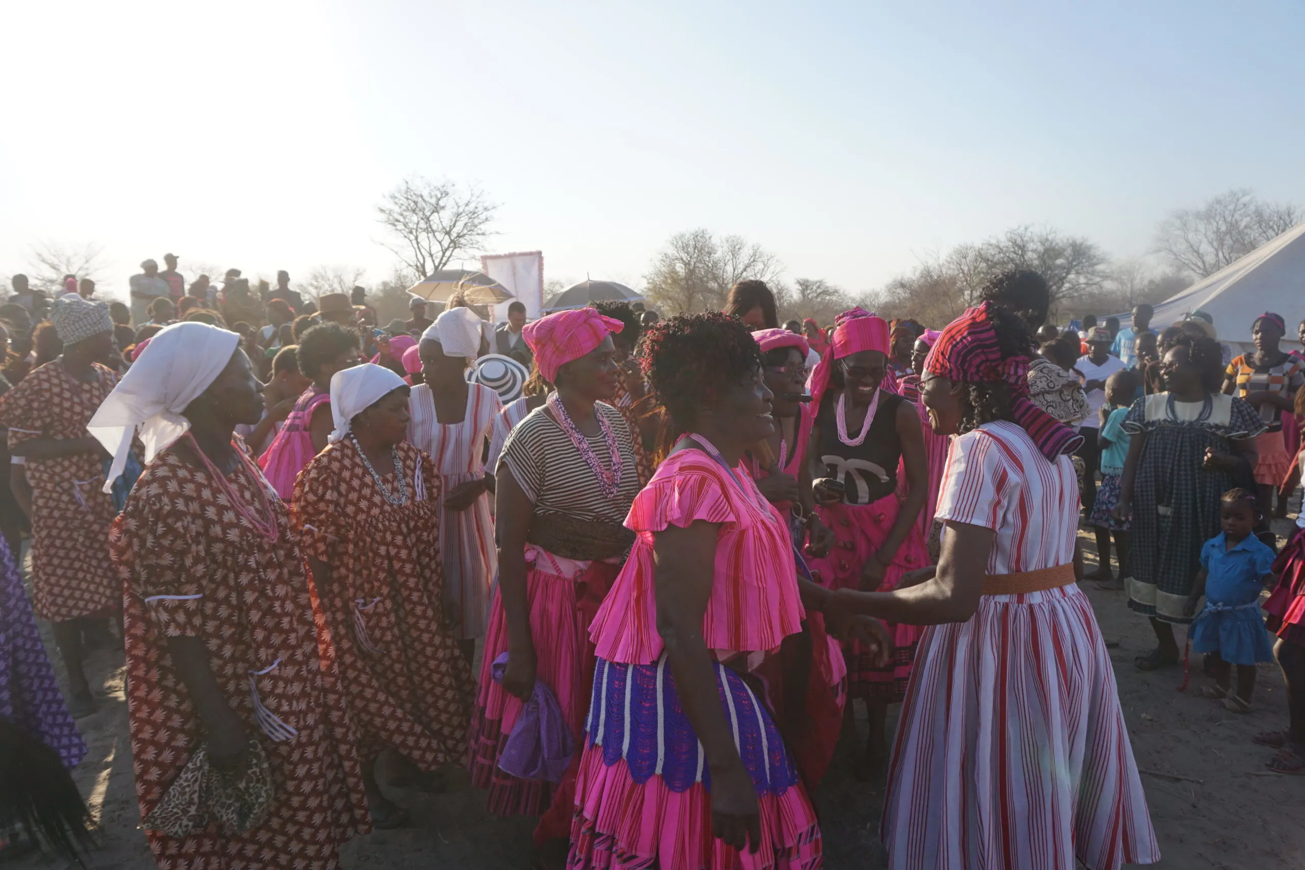 Eine Menschenmenge in Namibia versammelt sich zu einer traditionellen Hochzeit in einem Dorf. Die Menschen tragen bunte traditionelle Kleidung, die reich an Mustern und Farben ist. Frauen tragen kunstvoll gestaltete Kleider oder Röcke mit bunten Stoffen, während Männer traditionelle Gewänder oder Hemden mit bunten Mustern tragen. Die Kleidung spiegelt die kulturelle Vielfalt und Traditionen des Landes wider. Die Szene ist lebhaft und voller Energie, während die Gäste sich versammeln, um die feierliche Veranstaltung zu genießen.