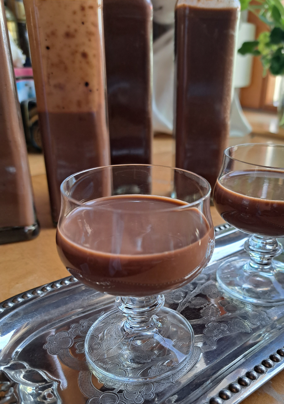 In einem Glas wird verführerischer Schokoeierlikör eingegossen, während im Hintergrund Flaschen mit weiterem Schokoeierlikör stehen.