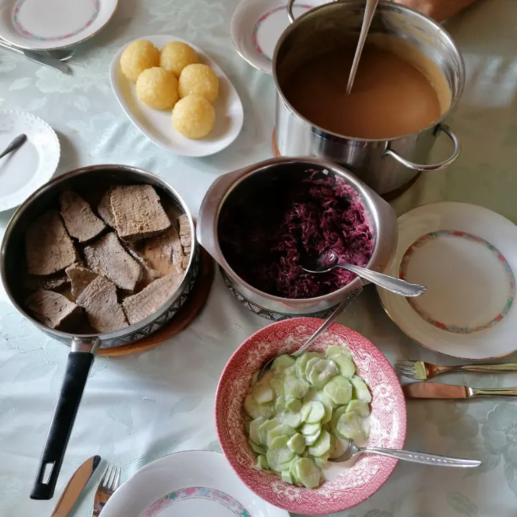 German food - A bountiful Sunday family meal set on the dining room table featuring a pot of sauerbraten sauce, tender sauerbraten meat (sour German braised beef), a platter of potato dumplings, a pot of Blaukraut (red cabbage), and a bowl of German-style cucumber salad.