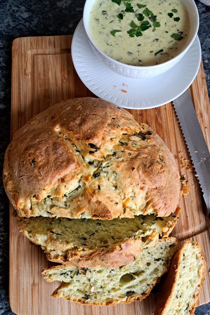 A cutting board with freshly baked wild garlic bread without yeast cut into slices, and a bowl of creamy wild garlic sauce next to it.