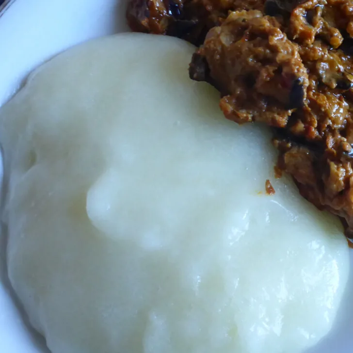 Einen Teller mit weißem Fufu aus Maniok, serviert mit einem afrikanischen Auberginensalat, bestehend aus u.a. Auberginen, Koriander, Cumin und Tomaten.