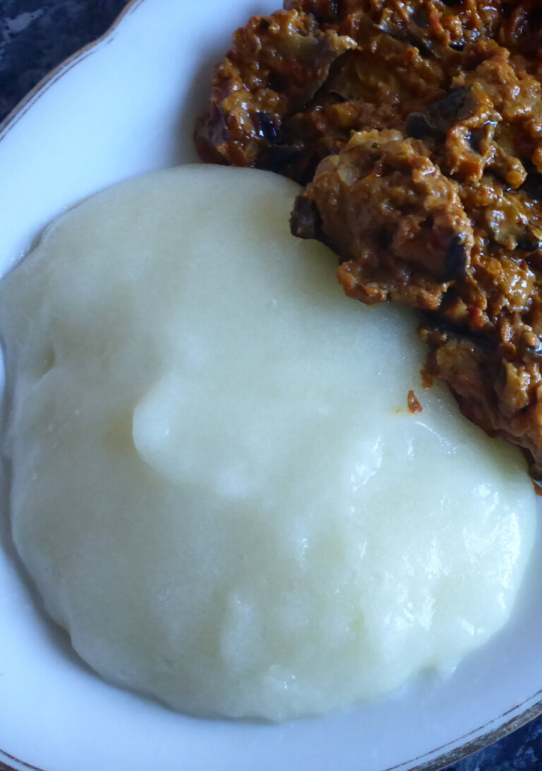 Einen Teller mit weißem Fufu aus Maniok, serviert mit einem afrikanischen Auberginensalat, bestehend aus u.a. Auberginen, Koriander, Cumin und Tomaten.