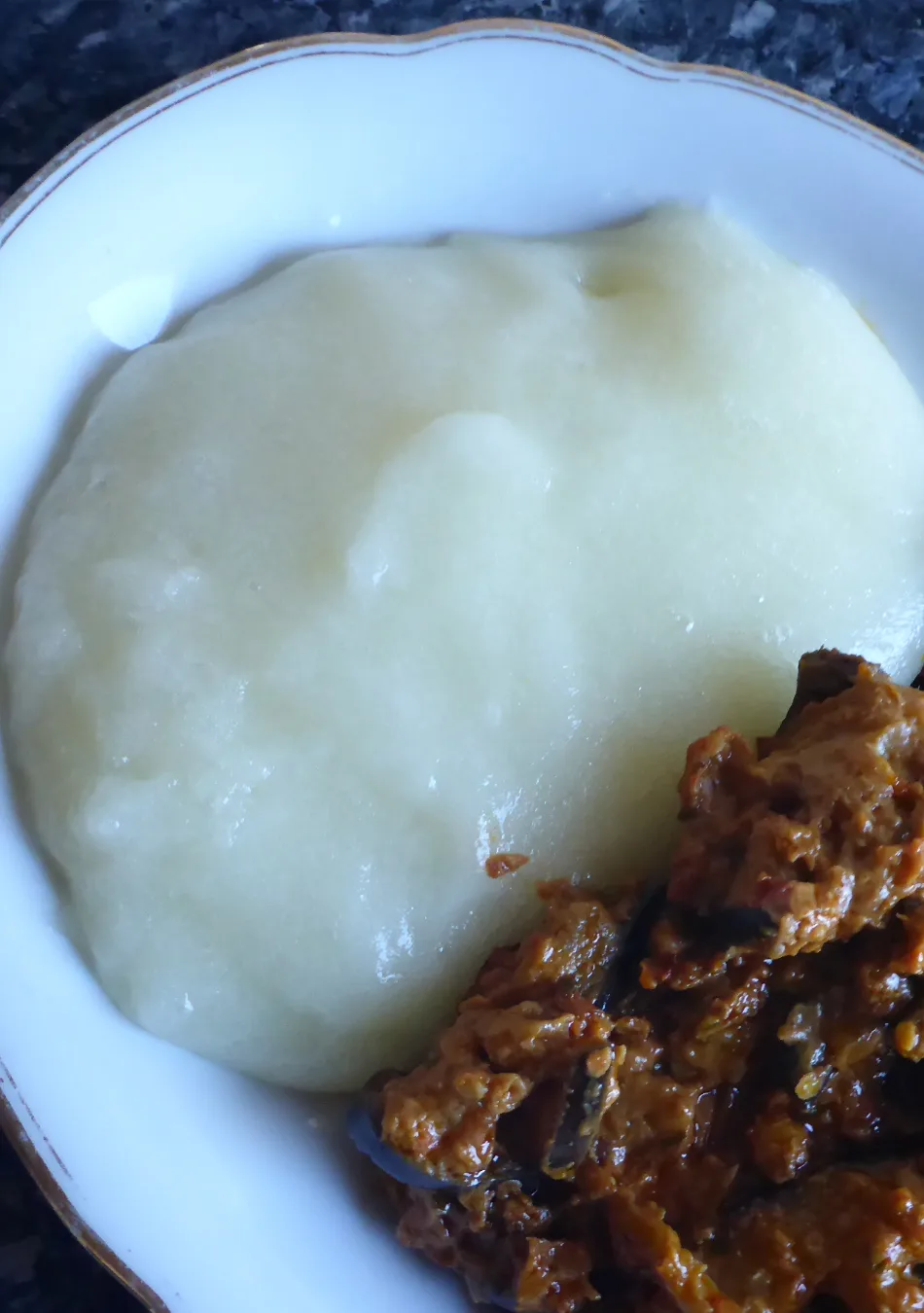 Einen Teller mit weißem Fufu aus Maniok, serviert mit einem afrikanischen Auberginensalat, bestehend aus u.a. Auberginen, Koriander, Cumin und Tomaten.