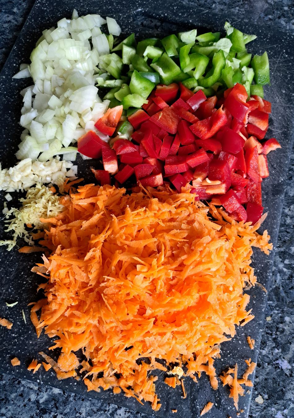 A cutting board with grated carrots, diced bell pepper (capsicum), grated ginger, minced onion, and garlic laid out in separate piles.