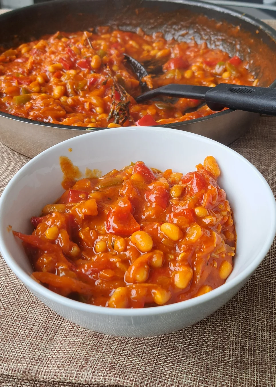 A pan of freshly cooked chakalaka, a South African vegetable relish, sitting on an outdoor balcony table. Next to the pan is a bowl of finished chakalaka, ready to be served. The relish is made with bell peppers, grated carrots, baked beans, garlic, fresh ginger, and onion, and can be enjoyed with a variety of foods for any meal of the day.