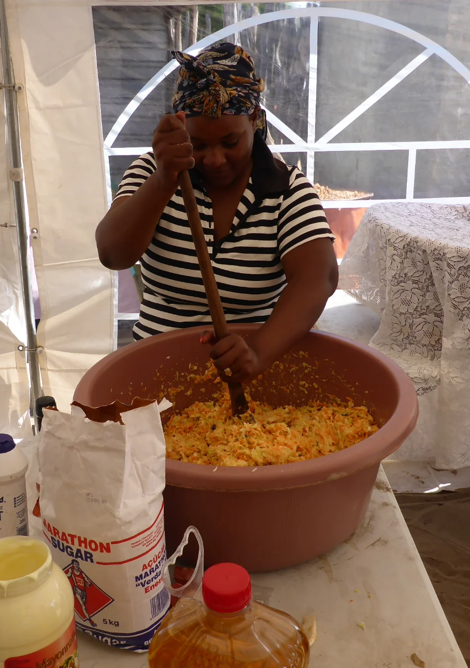 A woman inside a tent in a village in the north, energetically mixing coleslaw salad ingredients in a large bowl with a wooden spoon, surrounded by the rustic charm of a rural setting.
