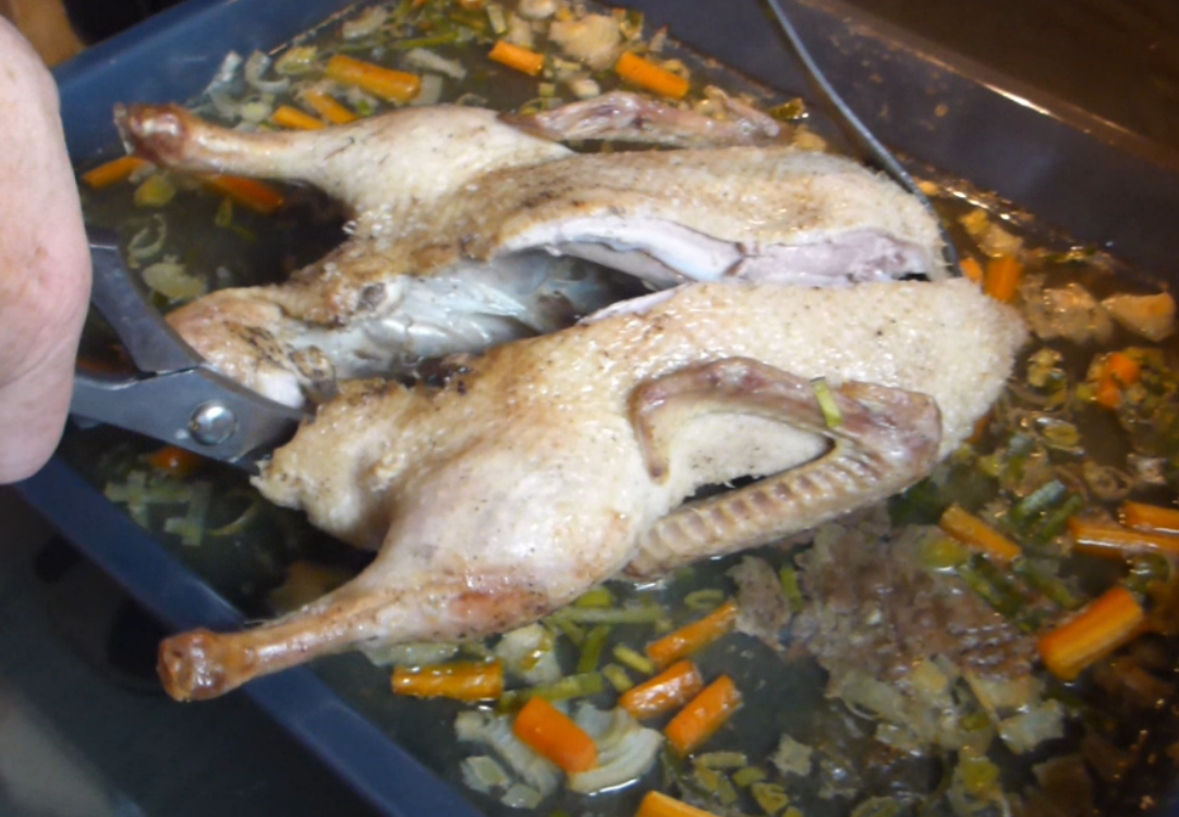 Ein Bild zeigt eine halb gekochte Ente auf einem Backblech, umgeben von Möhren und Lauch, während der Zubereitung im Backofen.