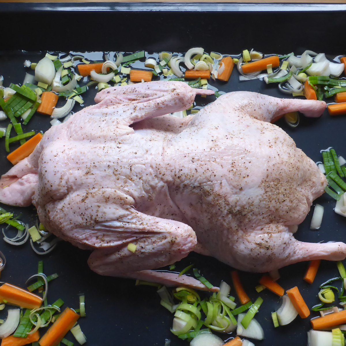Ein Bild zeigt ein Blech mit einer rohen Ente, umgeben von Möhren und Lauch, bereit zur Zubereitung im Backofen.