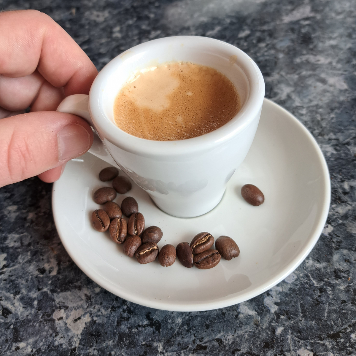 Frisch zubereiteten Espresso Siebträger in einer Espressotasse, umgeben von Kaffeebohnen auf einem Espresso-Unterteller. Eine Hand hält die Tasse und ist bereit, den Espresso zu genießen.