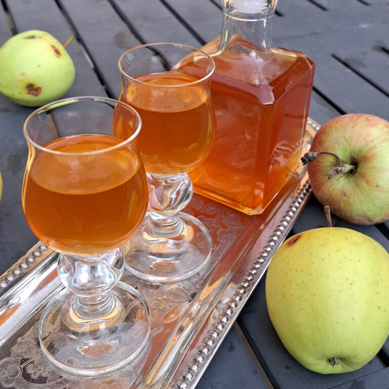Two glasses filled with insanely delicious homemade apple liqueur and a full bottle of the liqueur in the background.