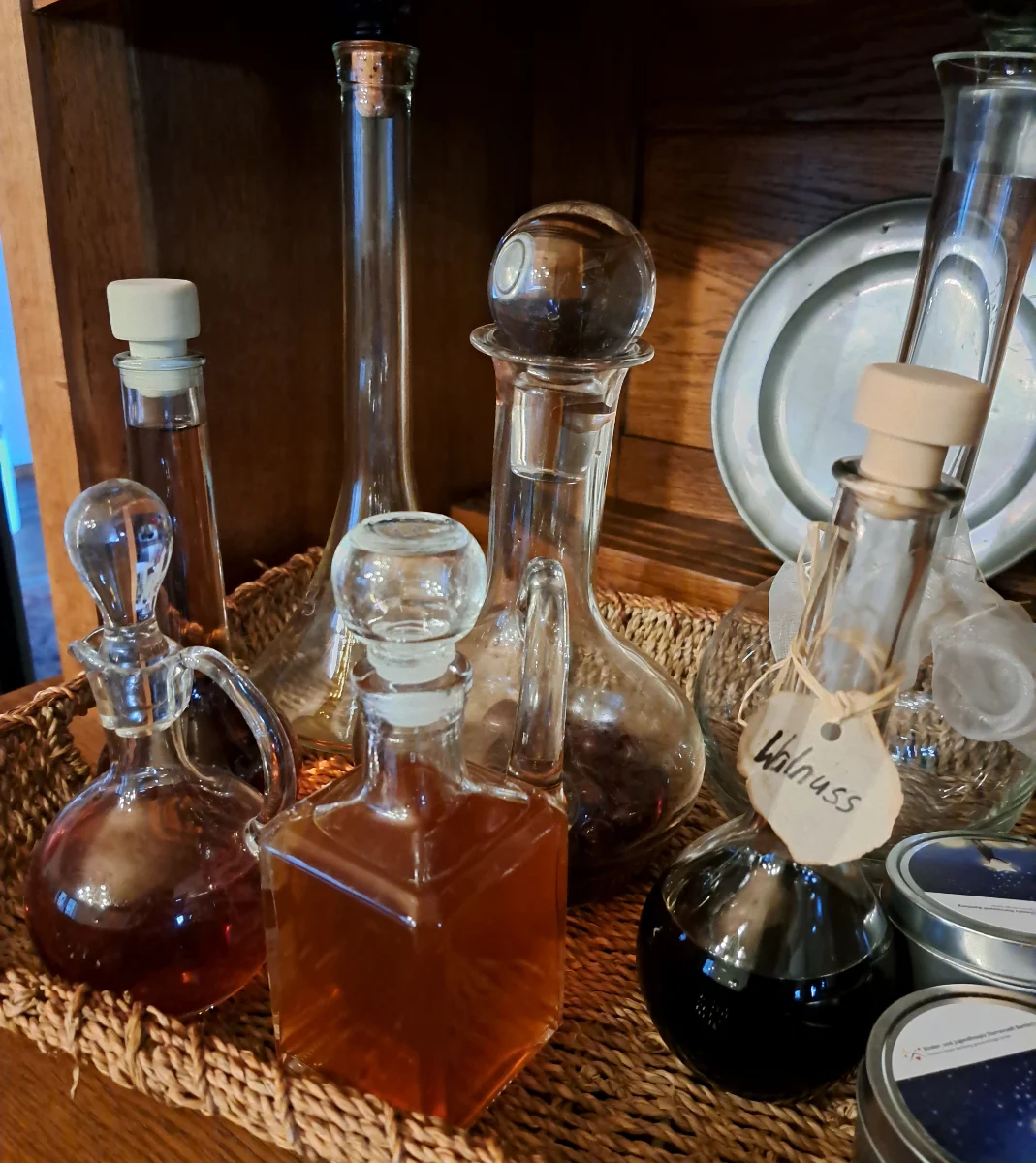 A photo of a variety of homemade liqueurs beautifully presented on a wooden serving board in a cozy living room, placed atop a cabinet or shelf. The liqueurs are arrayed in assorted bottles, creating an inviting liqueur board for guests to enjoy.