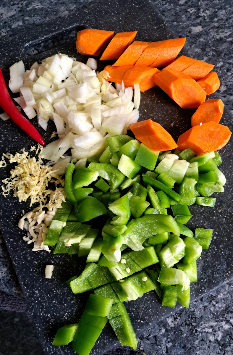 Fein geschnittenes Gemüse, darunter Paprika, Zwiebel, Knoblauch, Karotten und Ingwer, in Vorbereitung für die Zubereitung eines köstlichen Lammragouts auf afrikanische Art. Die bunten Zutaten versprechen ein reiches Geschmackserlebnis und eine aromatische kulinarische Reise.