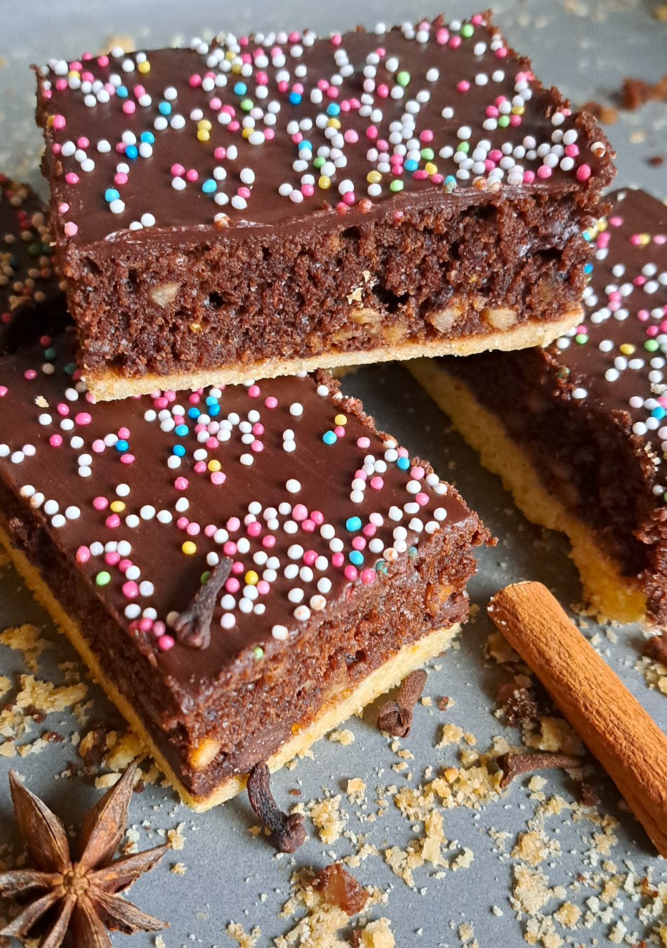 A visually appealing photograph featuring slices of German chocolate spice cake arranged in neat stacks, resembling gingerbread cake bars. The slices showcase a rich, moist texture and are garnished with whole spices such as star anise, cloves, and a cinnamon stick placed artfully beside them. The warm and inviting tones of the cake, coupled with the aromatic spices, create an enticing scene that captures the essence of traditional Gewürzkuchen.