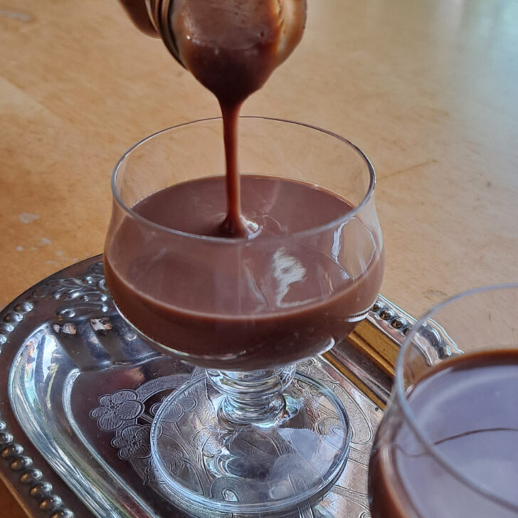 Pouring rich homemade chocolate Eierlikör from a bottle into a glass, capturing the luscious texture and enticing cocoa hues of this delightful liqueur.