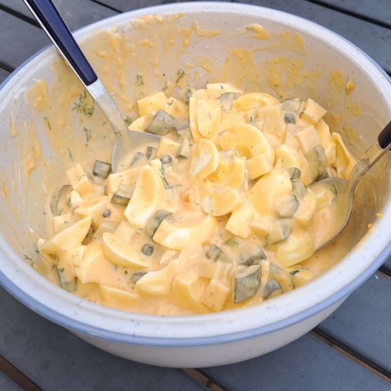 Ein Bild zeigt einen frisch zubereiteten Eiersalat in einer Schüssel. Die cremige Textur der Mayonnaise umhüllt die Eierstücke, während die knackigen Kapern, sauren Gurken und der frische Dill dem Salat eine köstliche Vielfalt verleihen. Perfekt für gesellige Anlässe oder als leichte Mahlzeit. Die appetitanregende Kombination aus frischen Zutaten macht diesen Eiersalat zu einem Genuss für alle Sinne.