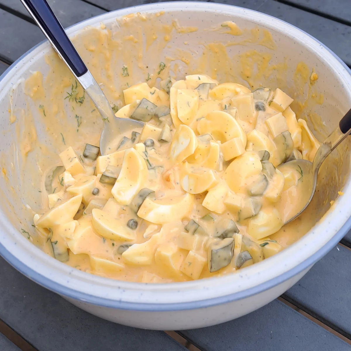Freshly made egg salad in a bowl, featuring hard-boiled eggs, capers, pickles, cheese, mustard, mayo, miracle whip, and dill.
