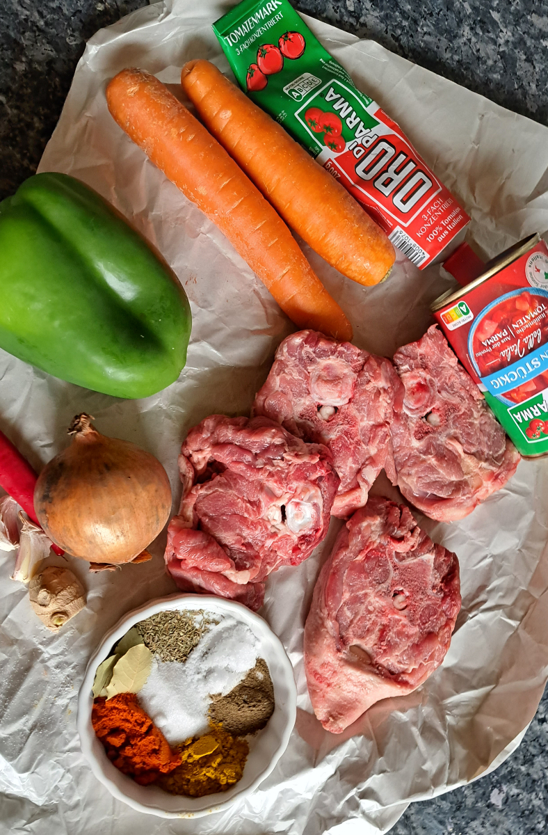 Image displaying a variety of fresh ingredients used to prepare lamb potjie, including lamb neck chops, fresh ginger, thyme, turmeric, bell pepper (capsicum), canned tomates, and garlic.