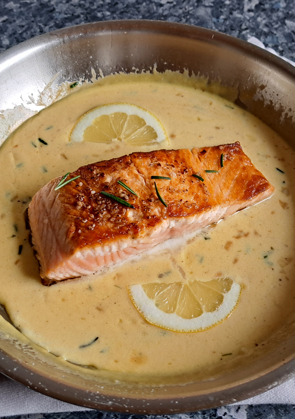 Pan-fried salmon immersed in a velvety, creamy orange sauce, adorned with vibrant sprigs of fresh rosemary and artfully arranged slices of lemon. The dish showcases a perfect blend of textures and flavors, creating a visually enticing and appetizing presentation.
