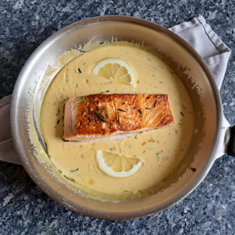 Pan-fried salmon immersed in a velvety, creamy orange sauce, adorned with vibrant sprigs of fresh rosemary and artfully arranged slices of lemon. The dish showcases a perfect blend of textures and flavors, creating a visually enticing and appetizing presentation.