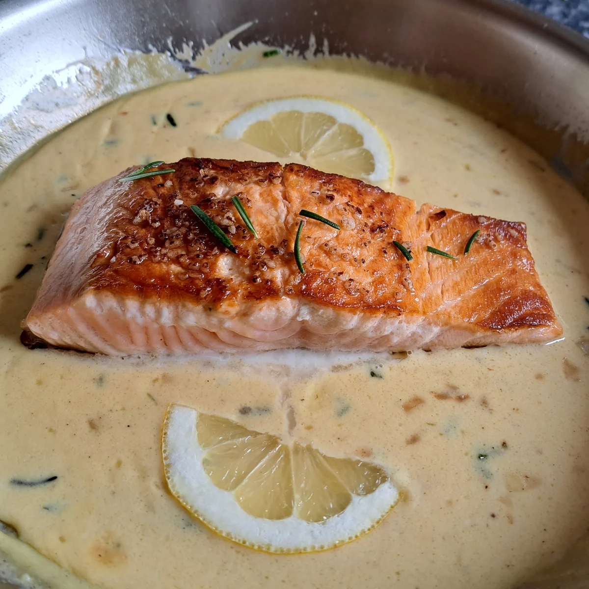 Pan-fried salmon fillet nestled in a rich and creamy orange sauce, with a vibrant blend of orange juice, mustard, and cranberry flavors, garnished with fresh rosemary and lemon slices. The salmon is perfectly seared, creating a visually appealing dish with a balance of colors and textures.