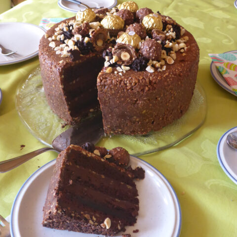 Eine beeindruckende Schoko-Nusstorte präsentiert sich, kunstvoll auf einem Tisch arrangiert und in perfekte Stücke geschnitten. Auf dem Teller liegt ein verlockendes Stück der Torte, die mit Ferrero Rocher Pralinen gekrönt ist. Die opulente Schichtung der Schokolade und Nusscreme wird durch die goldene Pralinenverzierung betont. Ein visuelles Festmahl, das den Genuss erahnen lässt.