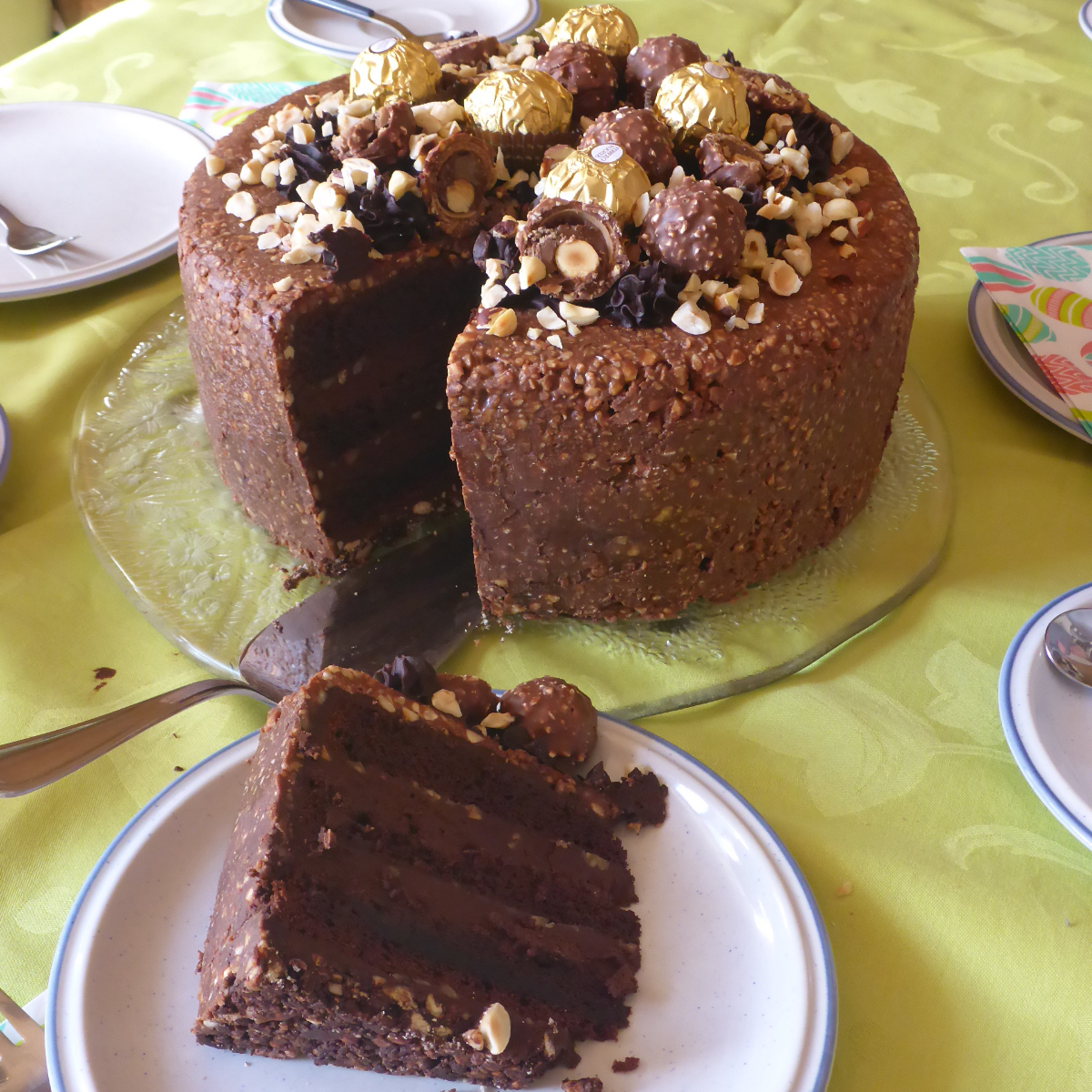 Eine beeindruckende Schoko-Nusstorte pr&auml;sentiert sich, kunstvoll auf einem Tisch arrangiert und in perfekte St&uuml;cke geschnitten. Auf dem Teller liegt ein verlockendes St&uuml;ck der Torte, die mit Ferrero Rocher Pralinen gekr&ouml;nt ist. Die opulente Schichtung der Schokolade und Nusscreme wird durch die goldene Pralinenverzierung betont. Ein visuelles Festmahl, das den Genuss erahnen l&auml;sst.
