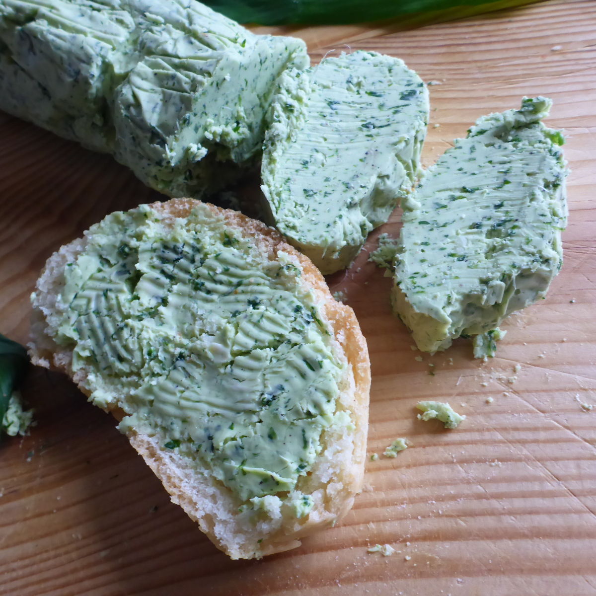 Ein Bild zeigt eine Bärlauchbutter-Rolle, die nach dem Einfrieren in Stücke geschnitten wurde. Neben den Stücken liegt eine Scheibe Baguette, auf die eine Portion Bärlauchbutter gestrichen ist.