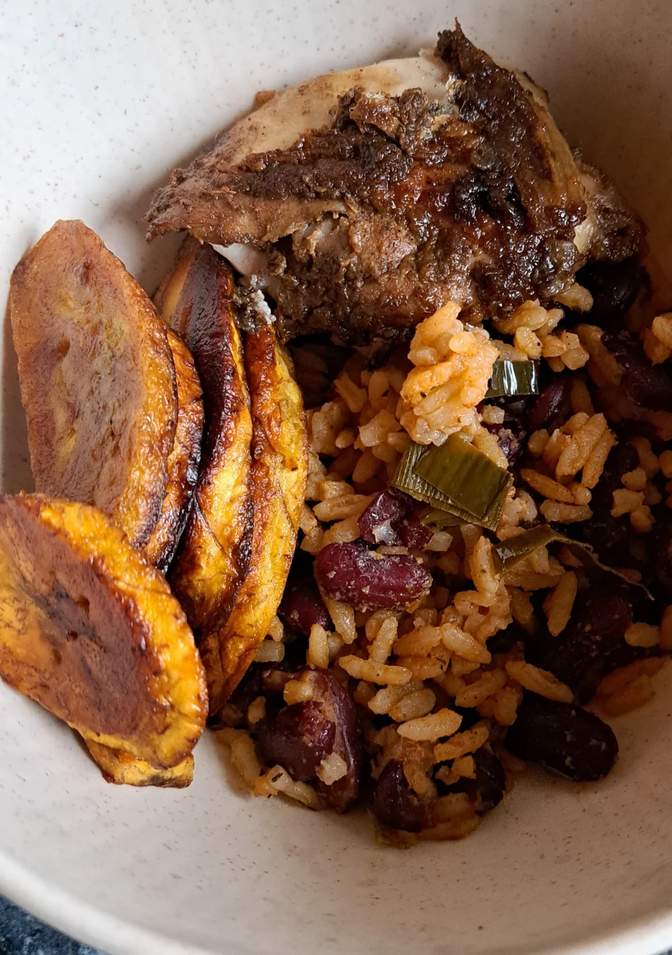 In einer Schüssel befindet sich jamaikanischer Reis mit Bohnen, dazu gebratene Kochbananen und jamaikanisches Hähnchen.