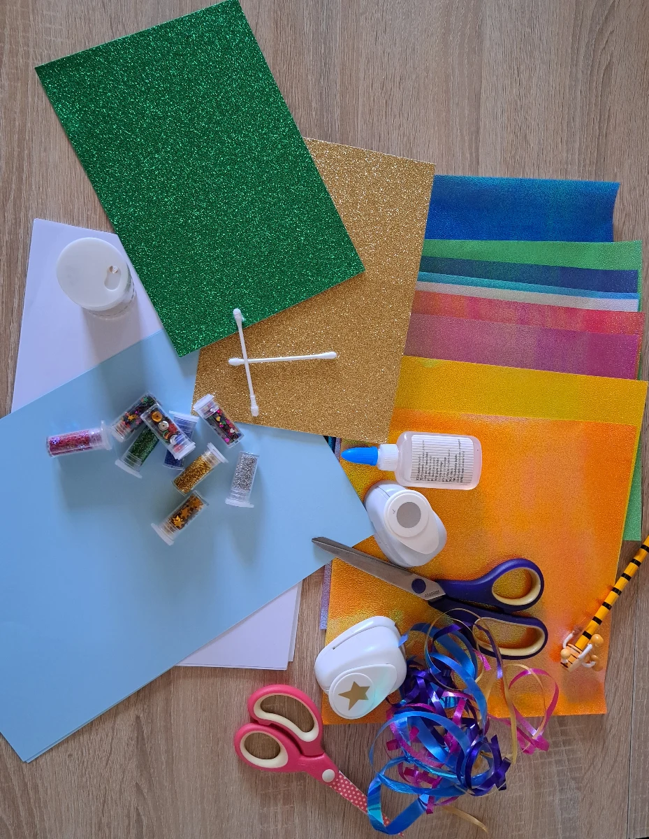 Das Bild zeigt die Materialien, die zur Herstellung der Meerjungfrau-Einladungskarten verwendet wurden. Zu sehen sind Glitzerpapier in verschiedenen Farben, ein Kreisstanzer, Scheren, Kleber, ein Kartonbogen, Bleistifte, sowie ausgeschnittene Fischschwanzflossen und gestanzte Glitzerkreise. Alle Materialien sind ordentlich auf einem Basteltisch angeordnet.