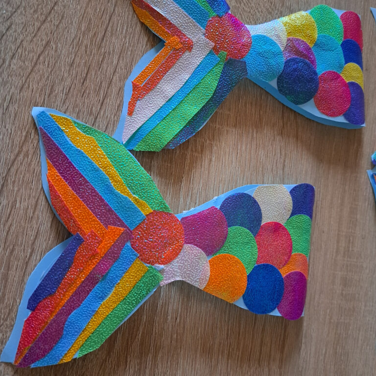 Die fertige Meerjungfrau-Einladungskarte in Form einer Fischschwanzflosse. Die Karte ist mit leuchtenden Schuppen aus Glitzerpapier und glänzenden Streifen verziert. Die Details sind sorgfältig platziert, und die Farben schimmern lebendig, wodurch die Karte einen funkelnden, festlichen Eindruck vermittelt.