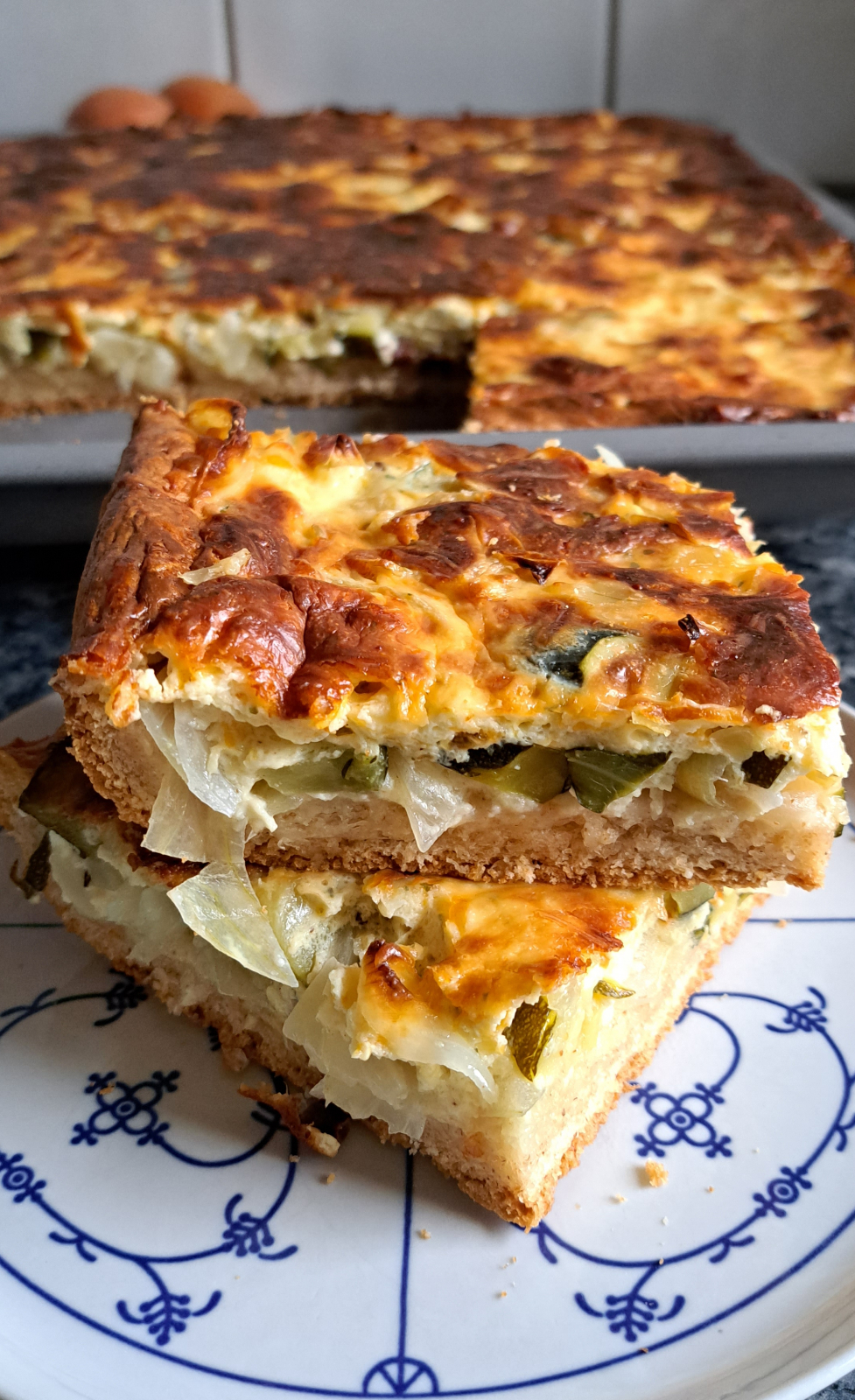 A plate with two slices of freshly baked German onion cake (Zwiebelkuchen) in the foreground, and the baking sheet with the remaining onion cake visible in the background where it was baked.