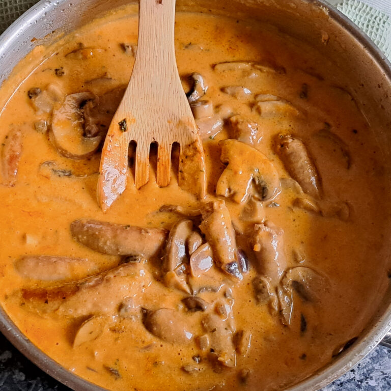 A deep pan filled with freshly cooked boerewors in a smooth and velvety creamy mushroom sauce. A wooden spoon rests inside the pan, stirring the luscious sauce.