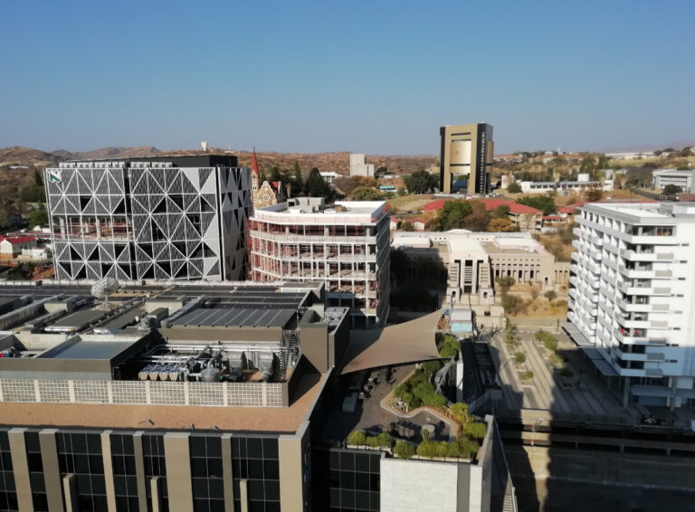 A breathtaking view of Windhoek from the top floor of Avani Hotel, showcasing the city's high-rise buildings, the historic Christuskirche, and the striking Independence Memorial Museum, built by North Korea. The cityscape blends modern and historical architecture against the scenic backdrop of the Namibian capital.