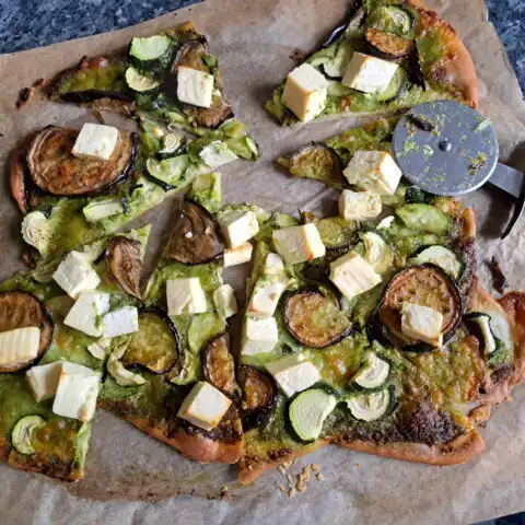 Frisch gebackene Pesto-Pizza mit Bärlauchpesto statt Tomatensoße, belegt mit Mozzarella, gerösteten Auberginen, rohen Zucchinischeiben und gebackenem Feta, knusprig gebacken auf einem Pizzastein, in Stücke geschnitten und servierbereit.