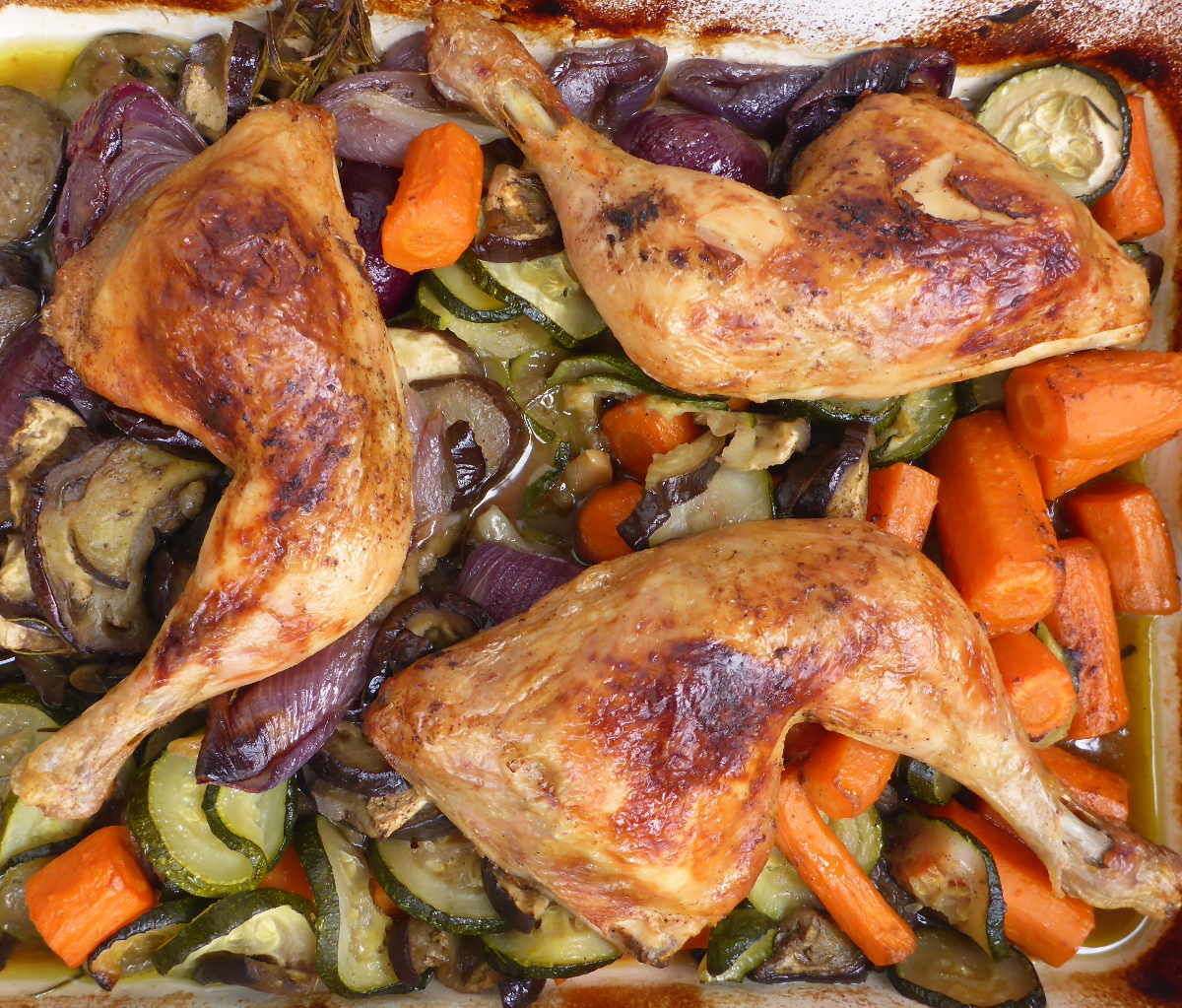 Frisch gebackene, knusprige Hähnchenschenkel aus dem Ofen auf einem Blech mit buntem Gemüse wie Zucchini, Karotten, Auberginen und Zwiebeln.